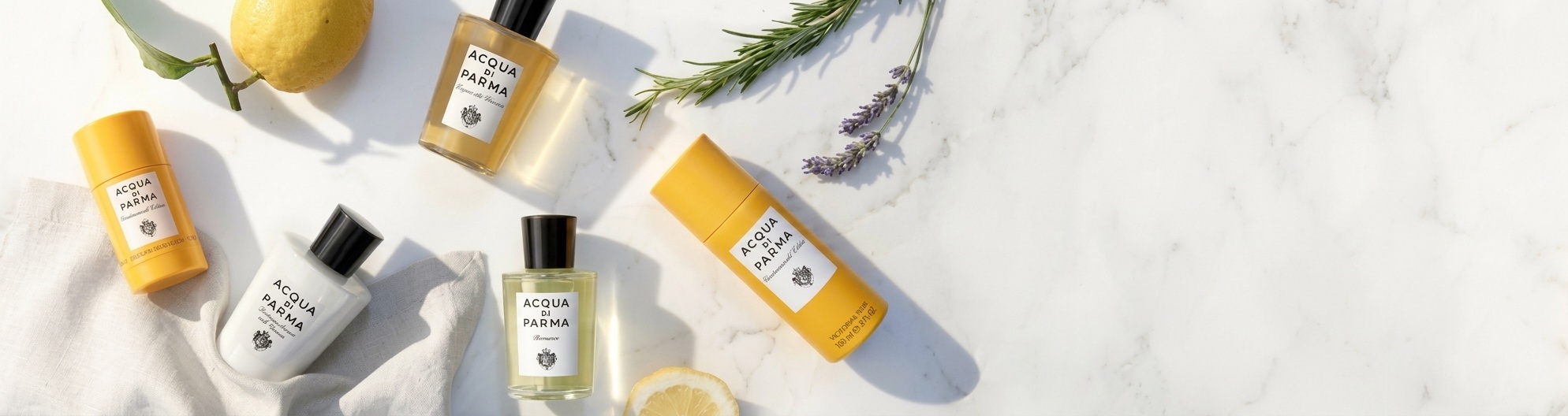 An overhead view of Acqua Di Parma perfume bottles, deodorants, fresh lemons, and lavender on a white marble background